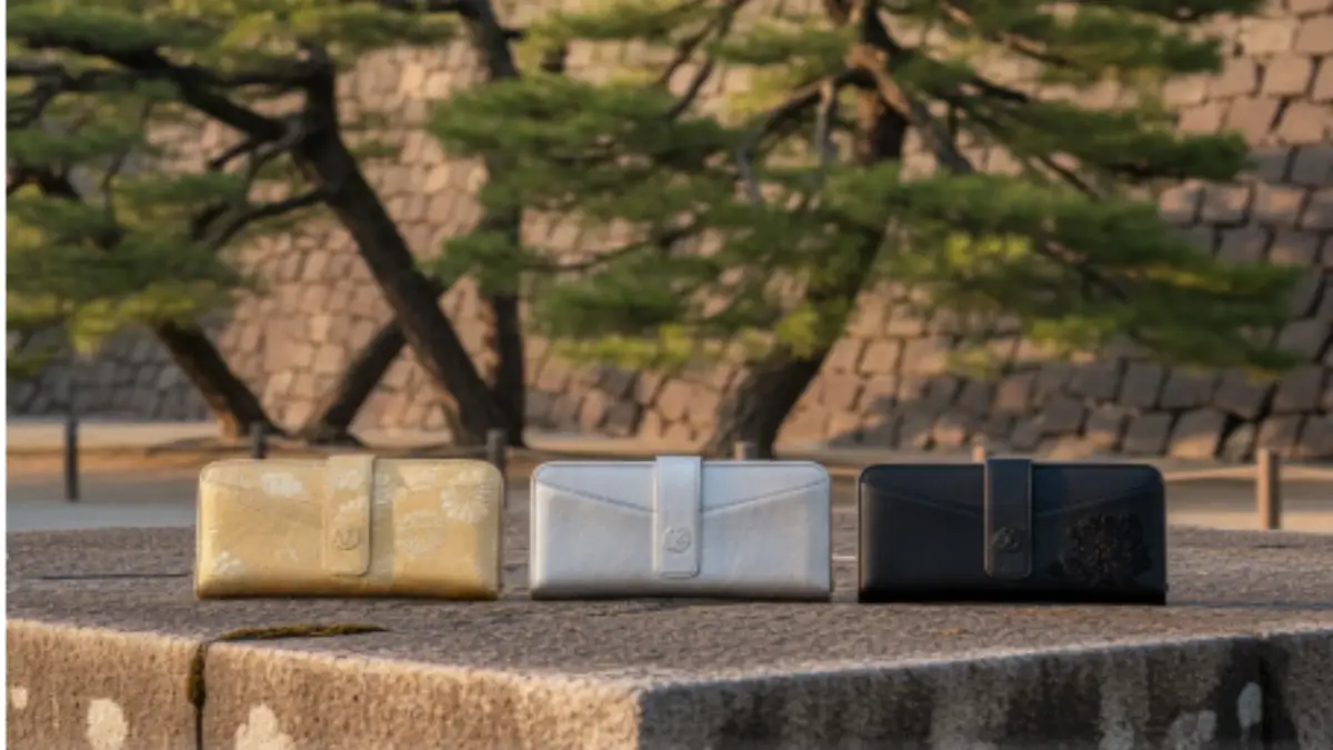 皇居限定の金・銀・黒の三色の長財布が、石の表面に並べられ、背景には宮殿の壁と松の木が見える、洗練された雰囲気の画像